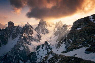 Rückenansicht eines nicht erkennbaren Touristen in Oberbekleidung, der auf Schnee steht und die Gipfel der Drei Zinnen gegen den wolkenverhangenen Himmel bei Sonnenuntergang in den Dolomiten, Italien, betrachtet - ADSF42145