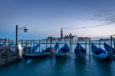 Row of traditional gondolas moored near pier in San Marco basin against San Giorgio Maggiore on island in cloudy evening in Venice, Italy - ADSF42151