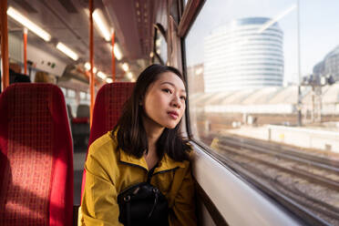 Junge asiatische Frau in Freizeitkleidung schaut aus dem Fenster, während sie tagsüber in London, Vereinigtes Königreich, Zug fährt - ADSF42155