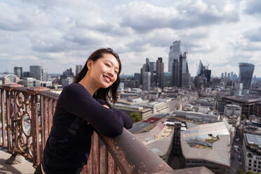 Seitenansicht einer fröhlichen asiatischen Frau, die sich auf ein Metallgeländer stützt und lächelnd vor Wolkenkratzern und bewölktem Himmel am Tag in London, Vereinigtes Königreich, wegschaut - ADSF42156