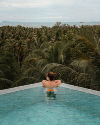 Back view of unrecognizable female tourist standing in corner of pool and looking around at lush green tropical plants in resort - ADSF42164