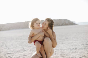 Lächelnde Mutter, die ihren Sohn umarmt, der am Strand kniet und seinen Urlaub genießt - MDOF00370
