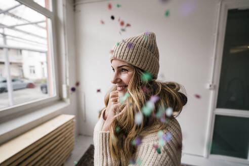 Happy blond woman wearing knit hat standing amidst falling confetti at home - JOSEF15527