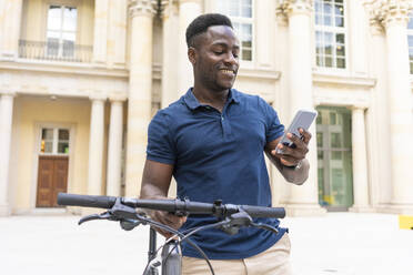 Smiling man using smart phone holding bicycle in front of building - TAMF03789