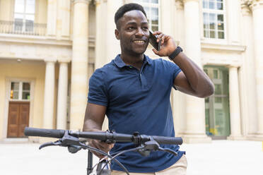 Smiling man talking on phone standing with bicycle in front of building - TAMF03790