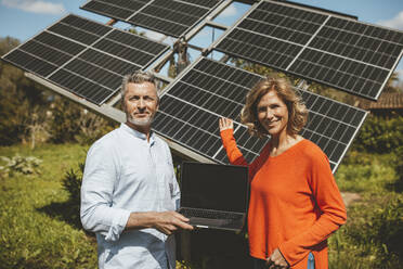 Glückliche reife Frau gestikuliert vor einem Mann mit Laptop vor einem Solarpanel - JOSEF15556