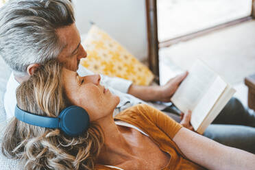 Ältere Frau mit kabellosen Kopfhörern sitzt neben einem Mann, der zu Hause ein Buch liest - JOSEF15563