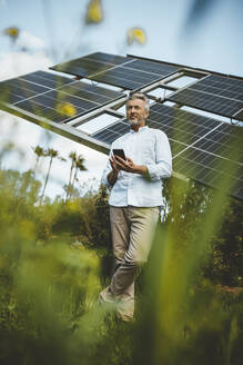 Älterer Mann lehnt sich mit seinem Smartphone im Garten an Sonnenkollektoren - JOSEF15566
