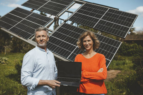 Lächelnder Mann und Frau mit Laptop vor Sonnenkollektoren an einem sonnigen Tag - JOSEF15586