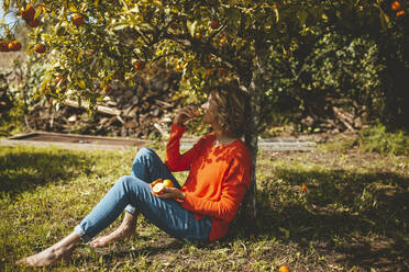 Mature woman eating orange fruit sitting by tree - JOSEF15595
