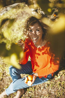 Glückliche Frau sitzt mit Orangenfrüchten unter einem Baum - JOSEF15598