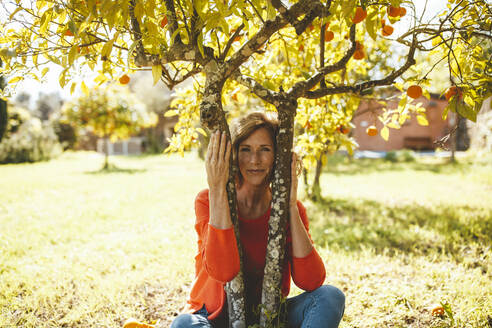 Lächelnde reife Frau sitzt unter einem Orangenfruchtbaum - JOSEF15603