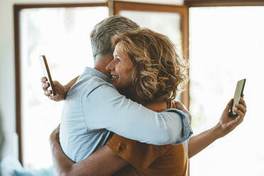 Mature couple with smart phones embracing each other at home - JOSEF15614