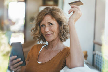 Lächelnde reife Frau sitzt mit Kreditkarte und Smartphone zu Hause - JOSEF15622