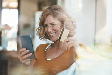 Happy mature woman sitting with credit card and using mobile phone - JOSEF15623