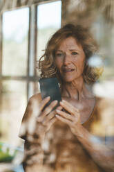 Lächelnde reife Frau, die ein Smartphone benutzt, gesehen durch ein Glasfenster - JOSEF15635