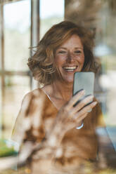 Happy woman using mobile phone seen through glass window - JOSEF15636