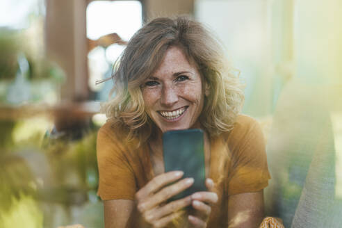 Glückliche Frau mit Smartphone, gesehen durch das Glas des Hauses - JOSEF15649