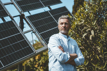 Thoughtful man with arms crossed in front of solar panels on sunny day - JOSEF15667