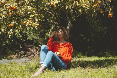Nachdenkliche Frau, die unter einem Baum sitzend Orangenfrüchte isst - JOSEF15691