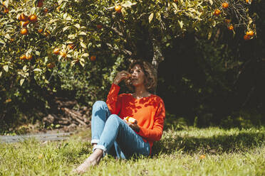 Thoughtful woman eating orange fruit sitting under tree - JOSEF15691