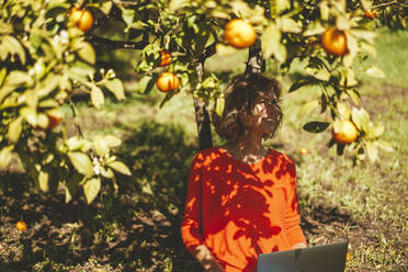 Kontemplative Frau sitzt mit Laptop unter einem Orangenbaum an einem sonnigen Tag - JOSEF15696