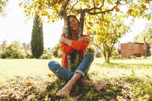 Reife Frau mit geschlossenen Augen umarmt orangefarbenen Obstbaum - JOSEF15700
