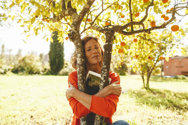 Woman with tablet PC hugging orange fruit tree in garden - JOSEF15701