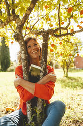 Happy mature woman with tablet PC hugging orange fruit tree - JOSEF15702