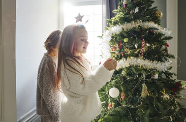 Glückliches Mädchen mit Bruder beim Schmücken des Weihnachtsbaums im Schlafzimmer zu Hause - MDOF00390