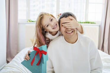 Glückliches Mädchen, das ein Weihnachtsgeschenk hält und die Augen des Vaters bedeckt, der zu Hause auf dem Bett sitzt - MDOF00392