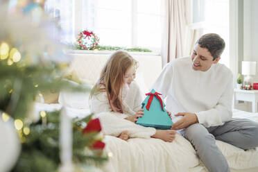 Glückliches Mädchen mit Weihnachtsgeschenk und Vater, der zu Hause auf dem Bett sitzt - MDOF00394