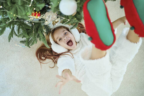 Cheerful girl lying on carpet by Christmas tree at home - MDOF00396