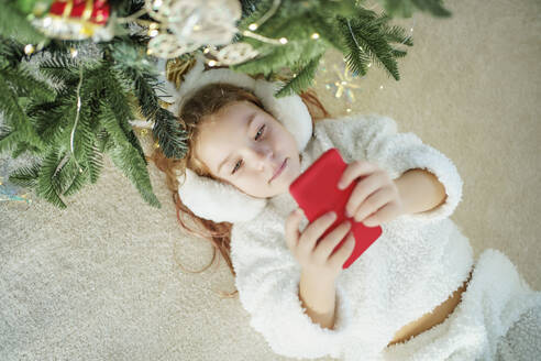 Girl lying on carpet by Christmas tree using smart phone at home - MDOF00397