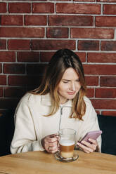 Frau, die ein Mobiltelefon benutzt und einen Cappuccino am Tisch im Café trinkt - VSNF00208