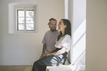 Glücklicher Hipster-Mann mit Freundin sitzt auf dem Tisch vor der Wand zu Hause - NDEF00312