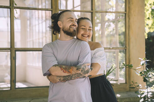 Happy woman embracing hipster boyfriend from behind in front of window - NDEF00337