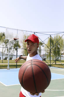 Junger Mann mit Mütze hält Basketball auf einem Sportplatz - SYEF00024