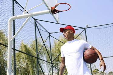 Junger Mann hält Basketball unter dem Arm an einem sonnigen Tag - SYEF00025