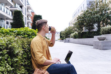 Young freelancer with laptop drinking coffee near plants - ASGF03162