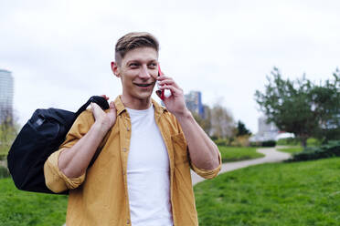 Junger Mann spricht mit Smartphone und trägt Tasche im Park - ASGF03176