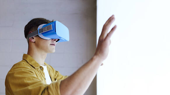 Junger Mann mit Virtual-Reality-Headset gestikuliert vor einer weißen Wand - ASGF03178