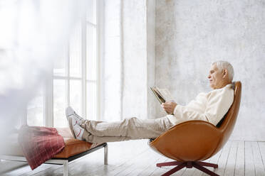 Senior man reading book sitting on chair at home - EYAF02388