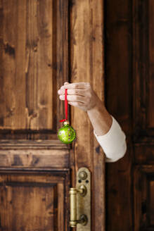 Eine Hand hält eine Weihnachtskugel vor einer Holztür - EYAF02401