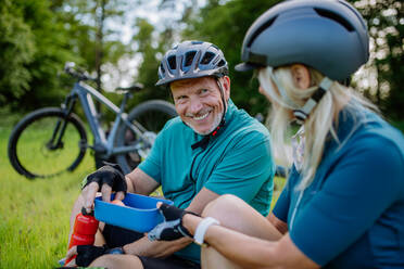 Ein aktives Seniorenpaar ruht sich nach einer Fahrradtour im Sommerpark aus, sitzt im Gras und isst eine Kleinigkeit. - HPIF03537