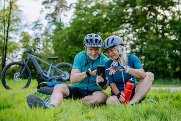 Ein aktives Seniorenpaar ruht sich nach einer Fahrradtour im Sommerpark aus, sitzt im Gras und benutzt sein Smartphone. - HPIF03541