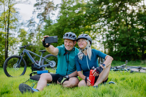 An active senior couple resting after bicycle ride at summer park, sitting on grass and taking slefie. - HPIF03542