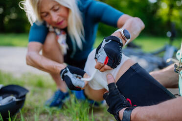 A senior woman is helping man after he fell off bicycle on the ground and injured his knee, in park in summer. - HPIF03550