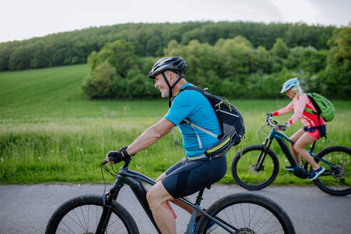 An active senior couple riding electric bicycles on path at summer park, healthy lifestyle concept. Side view. - HPIF03568