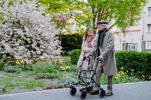 Ein älterer Mann mit Gehhilfe und erwachsener Tochter im Freien bei einem Spaziergang im Park. - HPIF03685